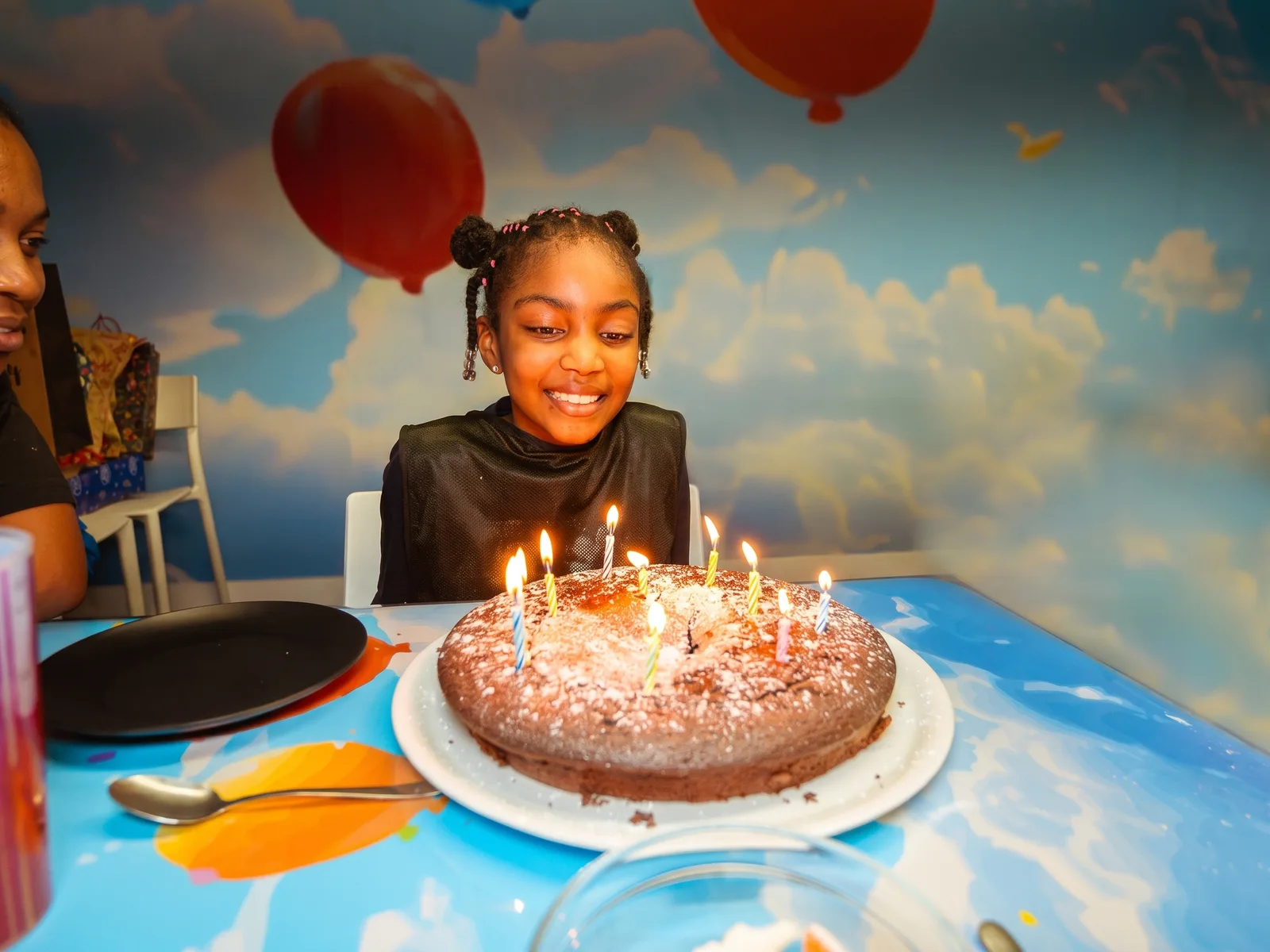 Une petite fille rayonnante devant son gâteau d'anniversaire aux bougies allumées, dans une salle NIKITO peinte en ciel et nuages
